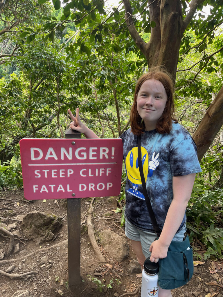 Cameron standing next to a sign that reads "Danger! Steep Cliff Fatal Drop" while smiling and making a peace sign on the Pipiwai Trial. 