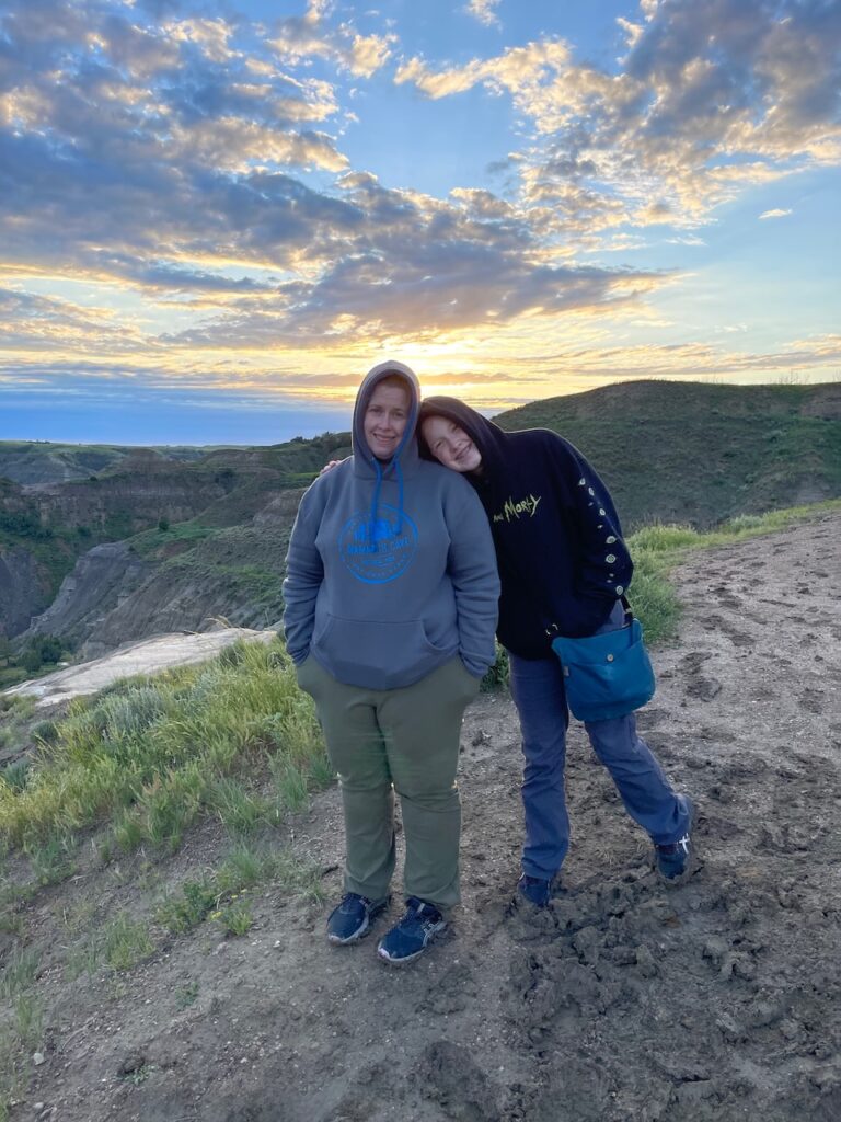 Cat and Cameron at a high view point in Theodore Roosevelt National Park, taken at sunset with clouds and color in the sky. It was rather cold and both women have their hoods up.