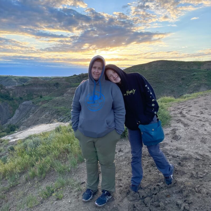 Cat and Cameron at a high view point in Theodore Roosevelt National Park, taken at sunset with clouds and color in the sky. It was rather cold and both women have their hoods up.