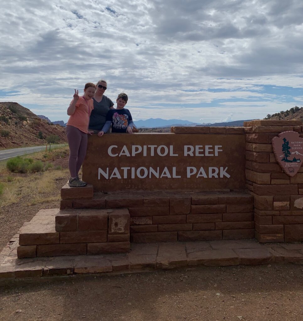 Cat, Cameron, and Collin standing on the park sign for Capitol Reef National Park with a big blue sky and clouds in the background.