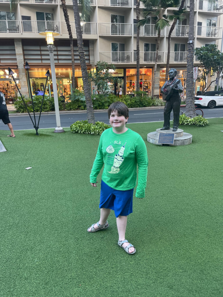 Collin standing in a city park in Waikiki in Honolulu - with shops and people all around.