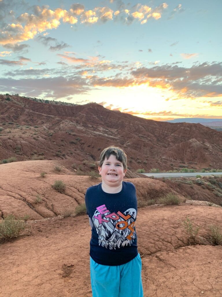 Cameron waiting with me for sunset on some rocks next to the Slickrock Divide. The sky is full of clouds and amazing colors and the winding  scenic drive road can be seen in the foreground.