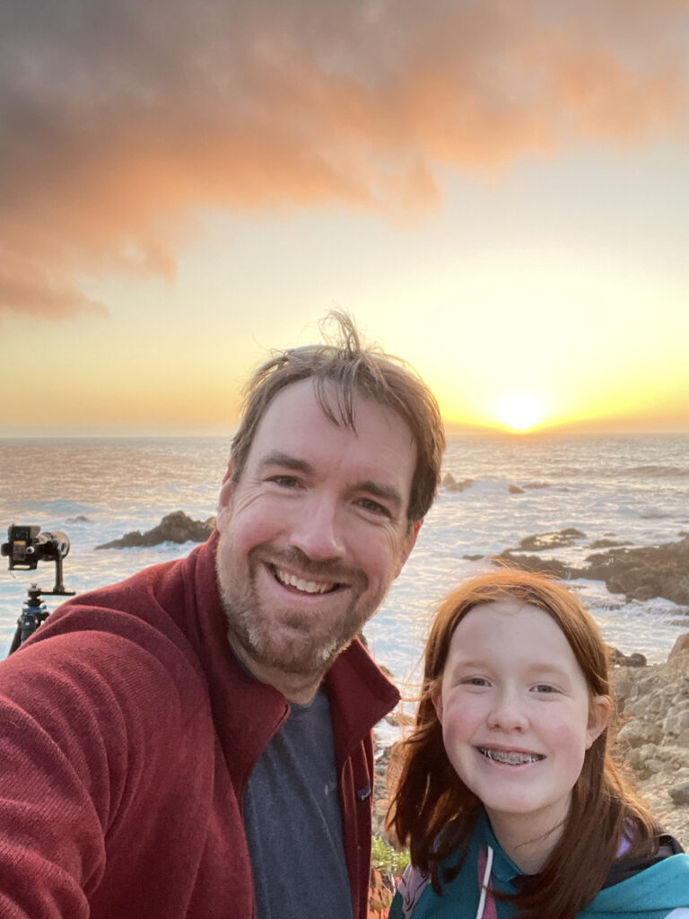 Cameron and myself posing for a photo with an amazing sunset over the pacific taking place behind us. Waves are crashing into the rocky shoreline and we are about halfway up the mountain side. 