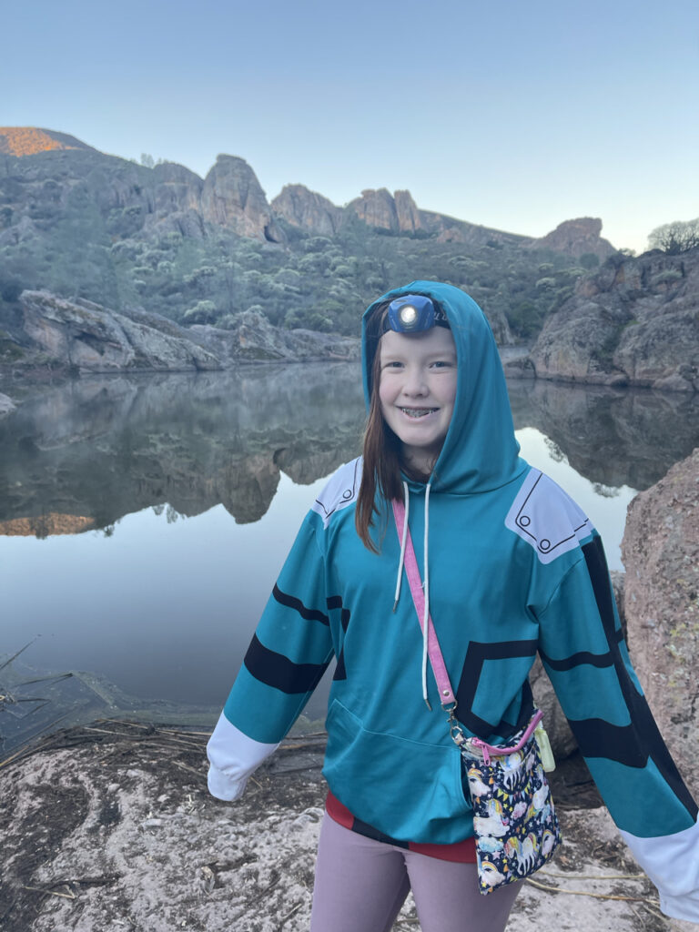 Cameron standing at the edge of the reservoir after hiking up the Bear Gulch trail. Still wearing her jacket and headlamp the sun is just hitting the peaks of the mountains across from her.