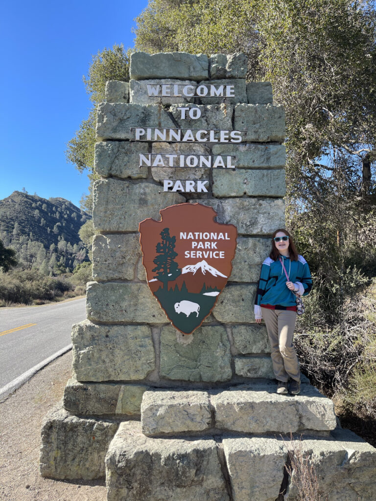 Cameron wearing a jacket and sunglasses standing on the stones next to the Pinnacles National Park Sign.