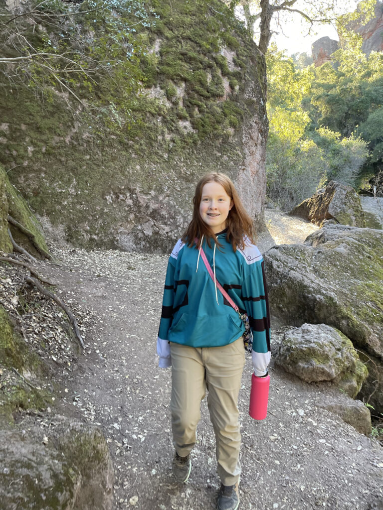 Cameron all smiles as she poses for a photo, holding my massive water bottle. Taken on the Rim Trail in Pinnacles National Park. Massive rocks, moss and trees surround the trial. 