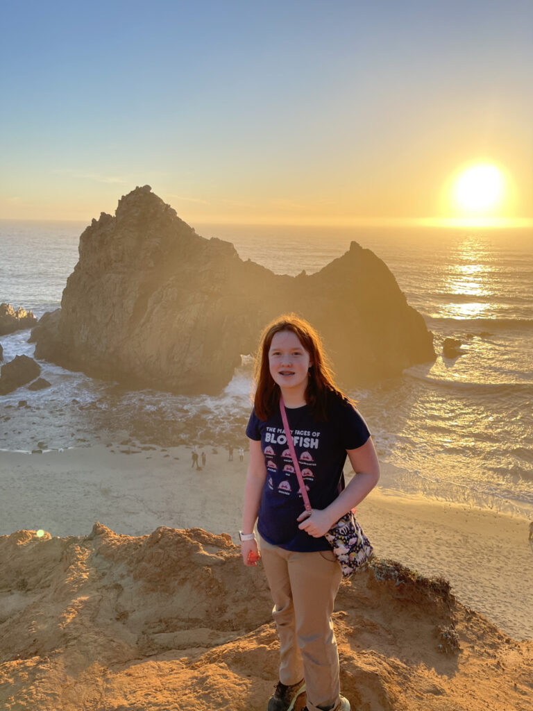 Cameron on top of the massive dune at Pfeiffer Beach, the sun is setting over the ocean behind us and making the cave in the rocks in the ocean glow.