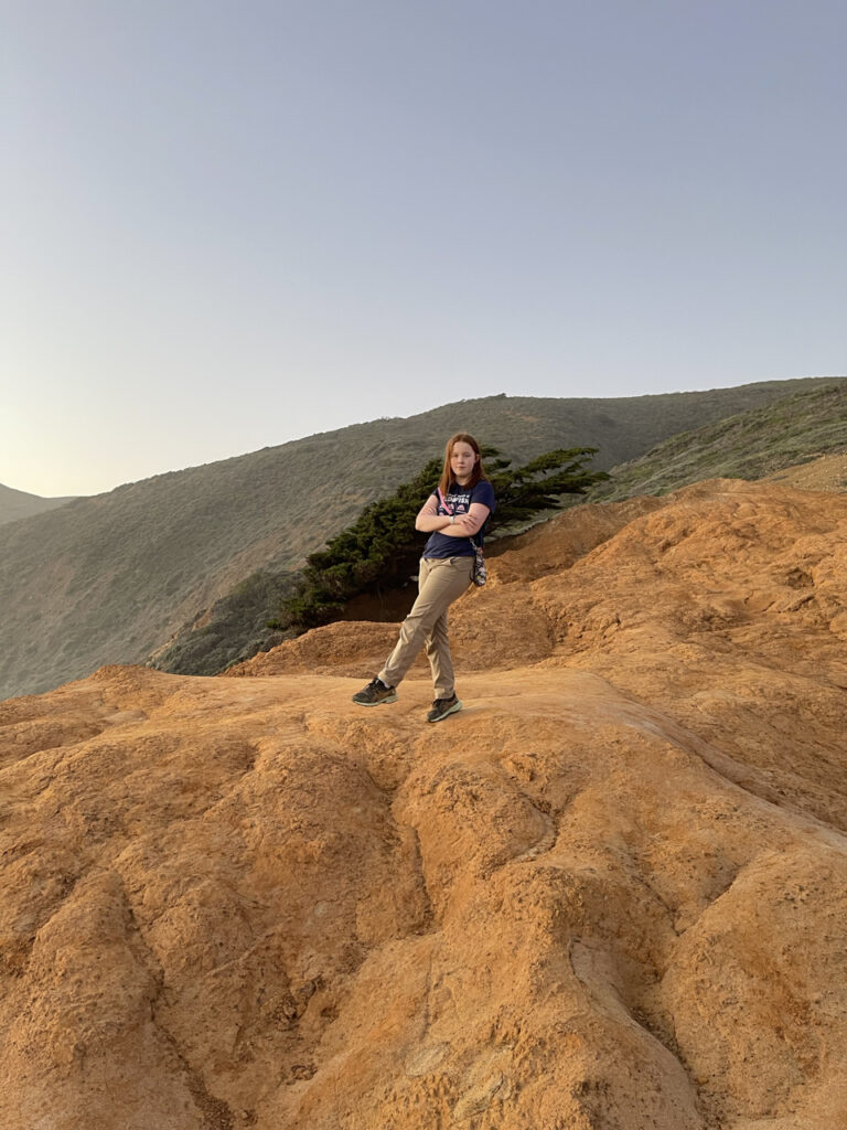 Cameron posing with her arms crossed and one one leg light with the last sunset light on Pfeiffer Beach in Big Sur.