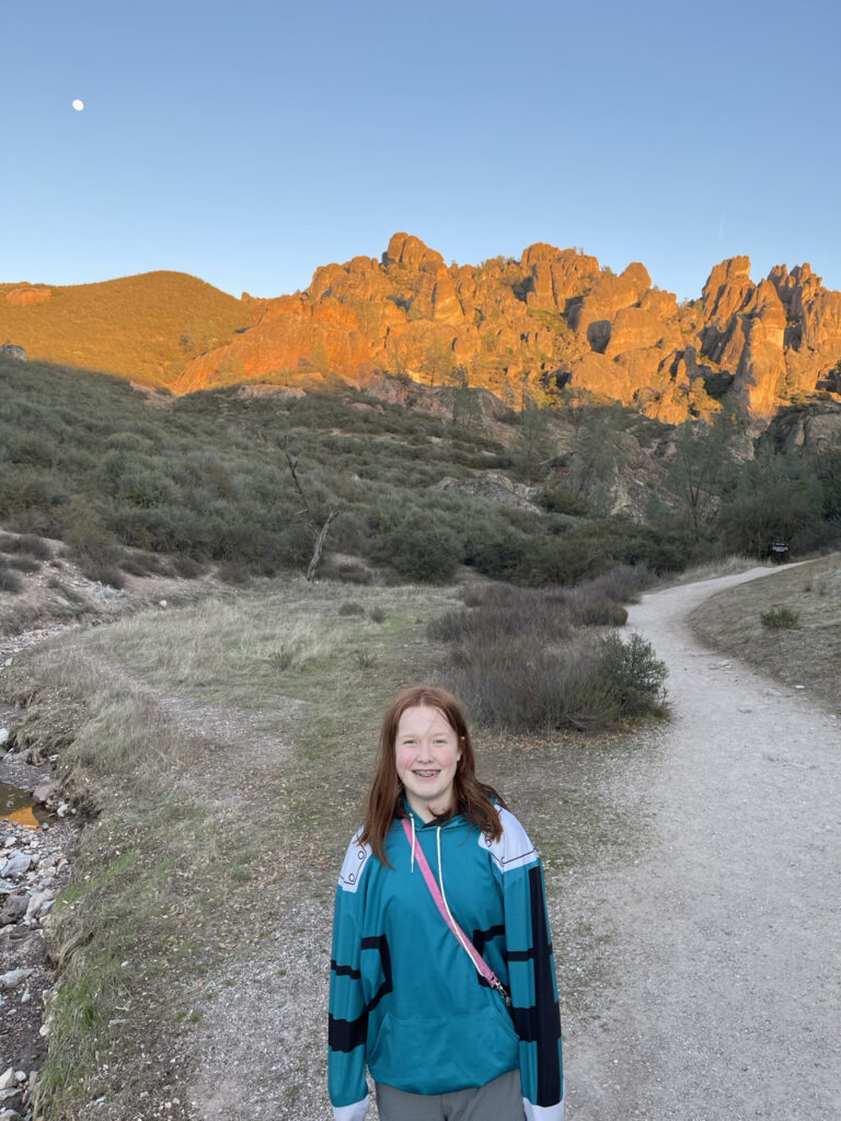 Cameron standing on the Juniper Canyon Trail, almost at sunset, with the Pinnacle Mountains behind her light up with the last light of the day. 