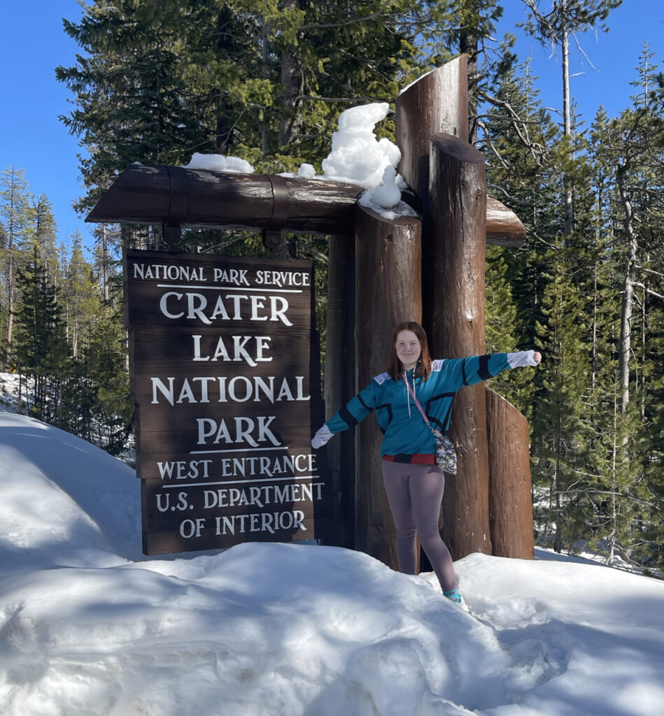 Cameron my red headed teen daughter standing on a massive pile of snow next to the Crater Lake National Park sign, that is also covered in snow. On a blue bird day in the middle of winter.