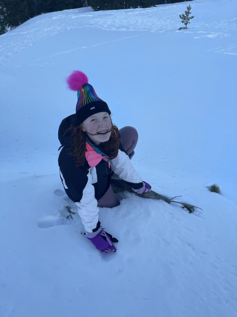 Cameron in a hat, gloves and snow jacket, laying in the snow with a massive smile on her face at Crater Lake National Park. The snow is so deep you can just see a tiny top to a tree next to her.