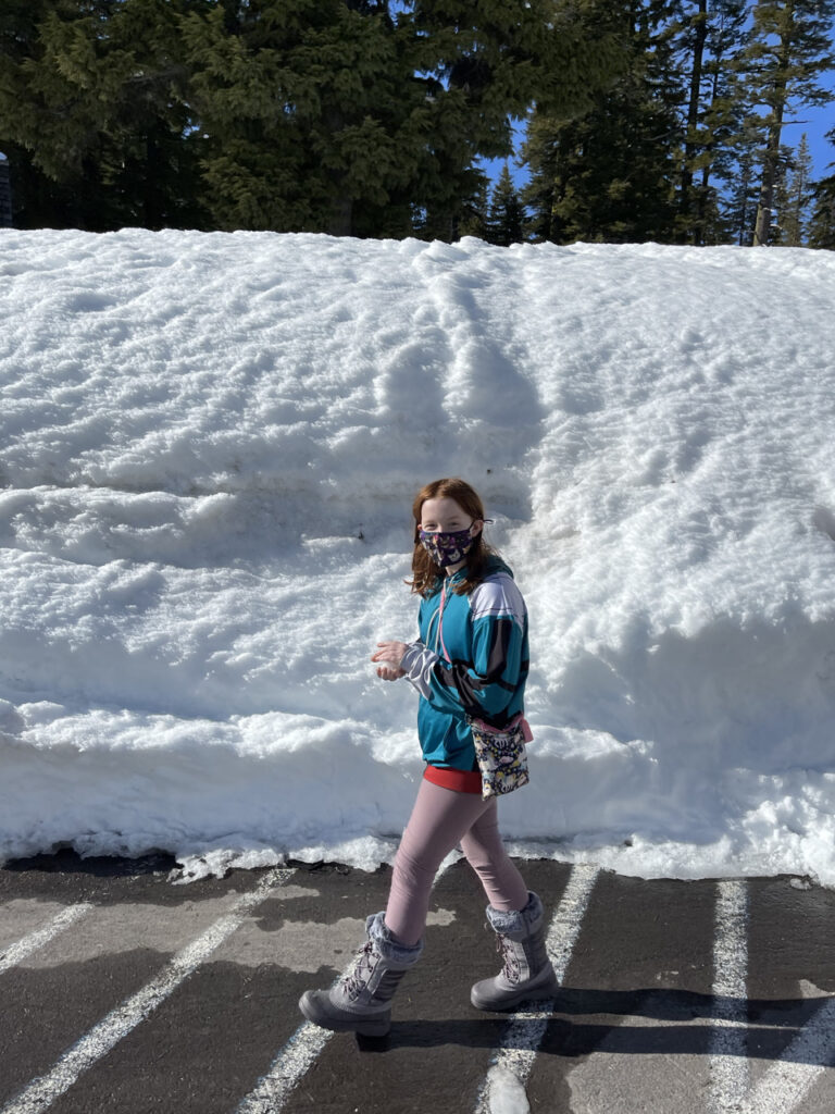 Cameron red headed teen wearing a jacket and mask walking in the Crater Lake Parking lot towards the visitors center and the snow bank behind her is twice as tall as she is.