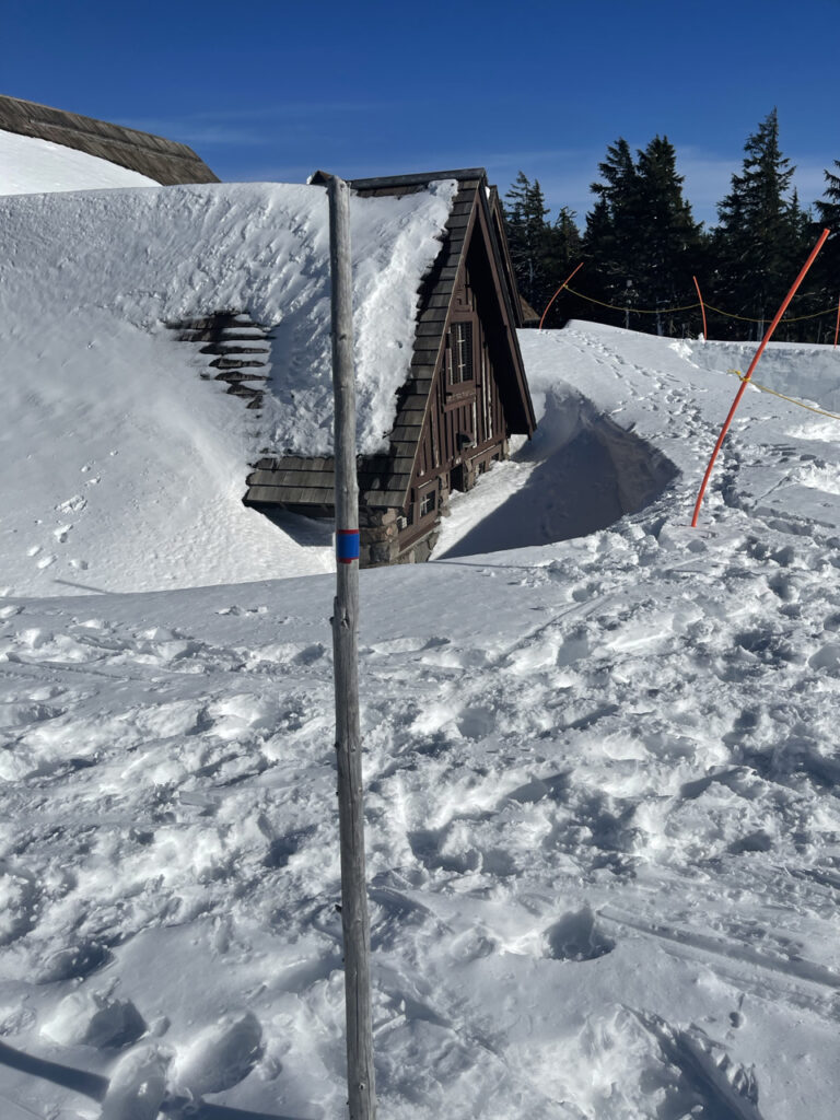 A picture of the Crater Lake Loge taken in winter with the snow almost covering the roof. One Second story window is still able to be seen. There are lots of tracks in the snow around the hotel.