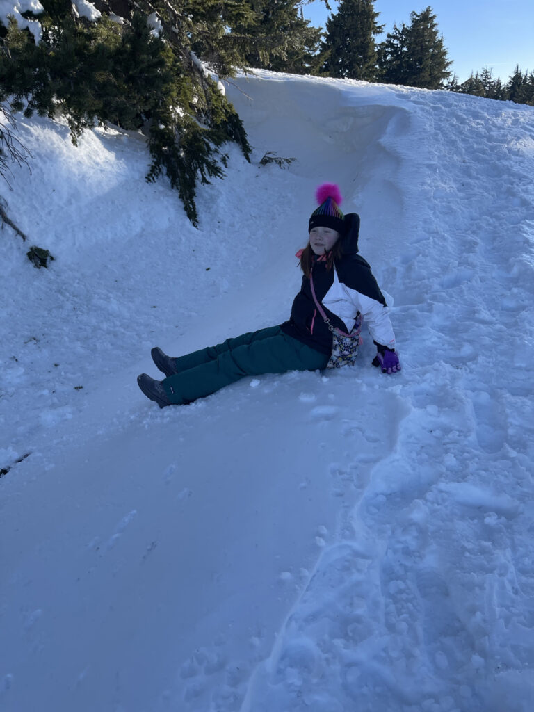 Cameron my teen daughter dressed in full snow gear sliding down a hill under a tree on a sunny winters day in Crater Lake.