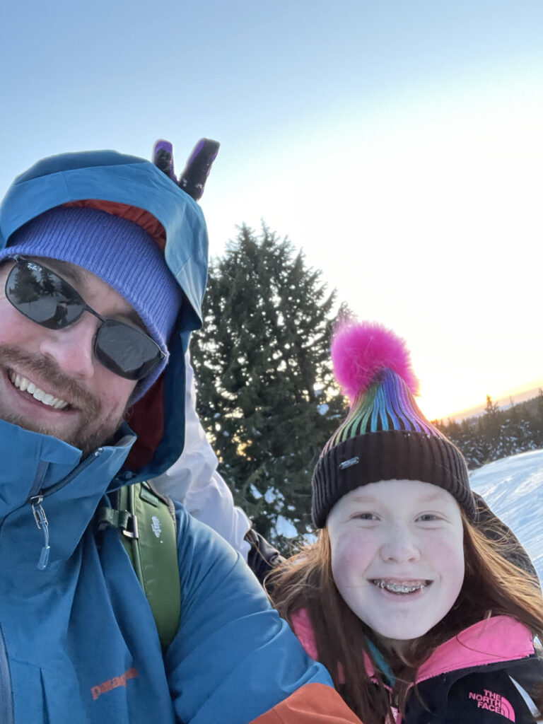 My daughter and myself standing on piles of snow in Crater Lake National Park. The lake is behind us and we are both fully dressed for cold winter days and I'm wearing sunglasses. She is smiling and making bunny ears over my head. 