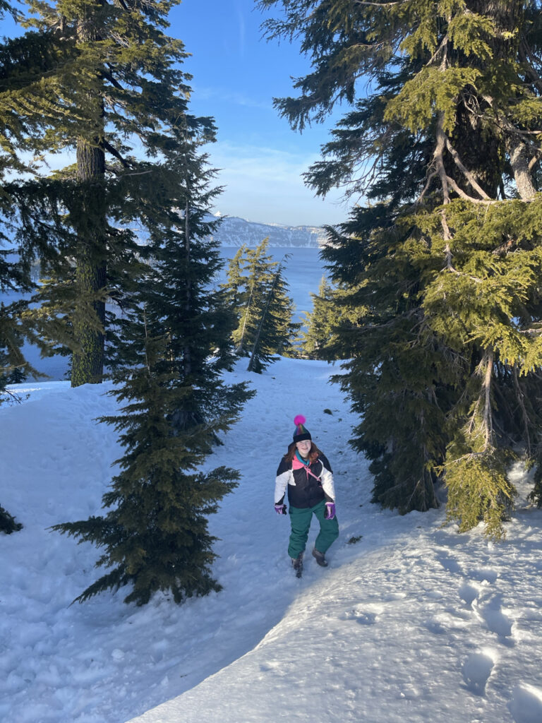 Cameron my red headed teen daughter posing for a photo in the shade on top of lots of snow at Crater Lake National Park. The trees around here are mostly buried  in snow and the lake is right behind her.