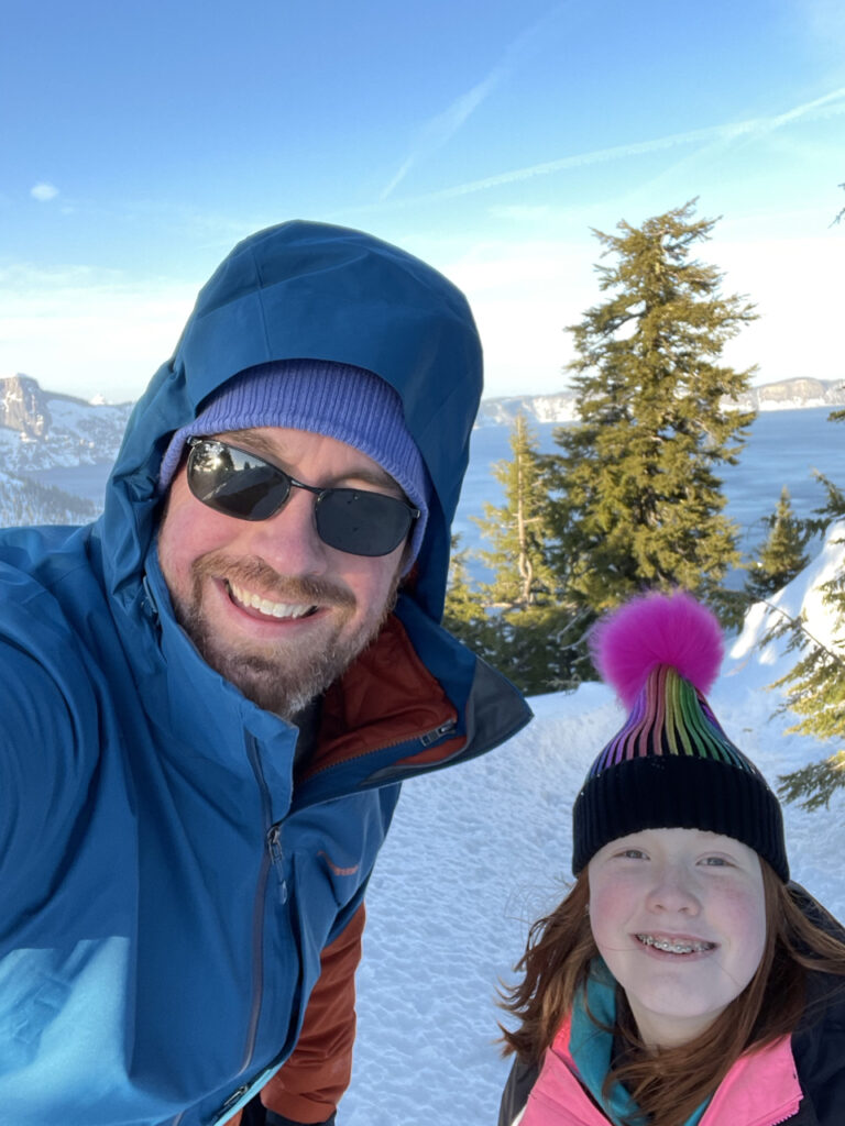 Myself with my daughter in full snow gear at Crater Lake National Park posing with the lake behind us on a blue bird sunny day in the mist of winter.