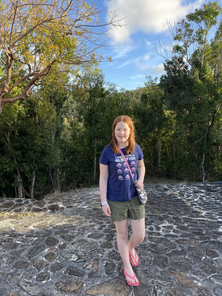 Cameron standing on the ground of the Catherineberg Sugar Mill Ruins, in shorts, a tshirt and flip-flops. The sun is hitting half her face and making her red hair glow.