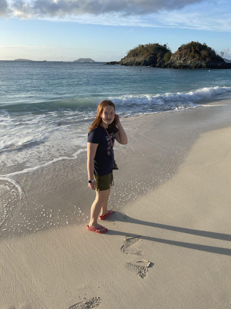 Cameron standing on the beach at the edge of the surf at Trunk Bay Beach in the US Virgin Islands. Wearing short and a t-shirt with flip-flops.