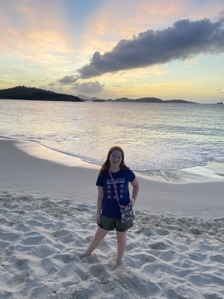 Cameron at Trunk Bay Beach in the US Virgin Islands. Standing in the sand at sunset with the ocean behind her. The rest of the island can be seen in the background along with red and yellow clouds from sunset.