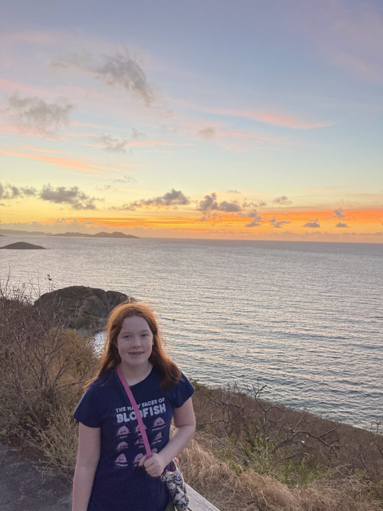 Cameron standing on a grassy cliff side, looking at the sunset over Drunk Bay, the clouds are orange and yellow.