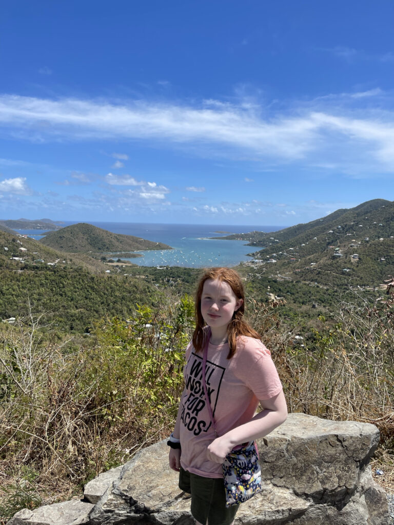 Cameron standing by a wall, near the top of the mountain on the side of the road in the Virgin Island. Looking down at tree lined valleys, an ocean bay full of boats and a skyline with wispy clouds.