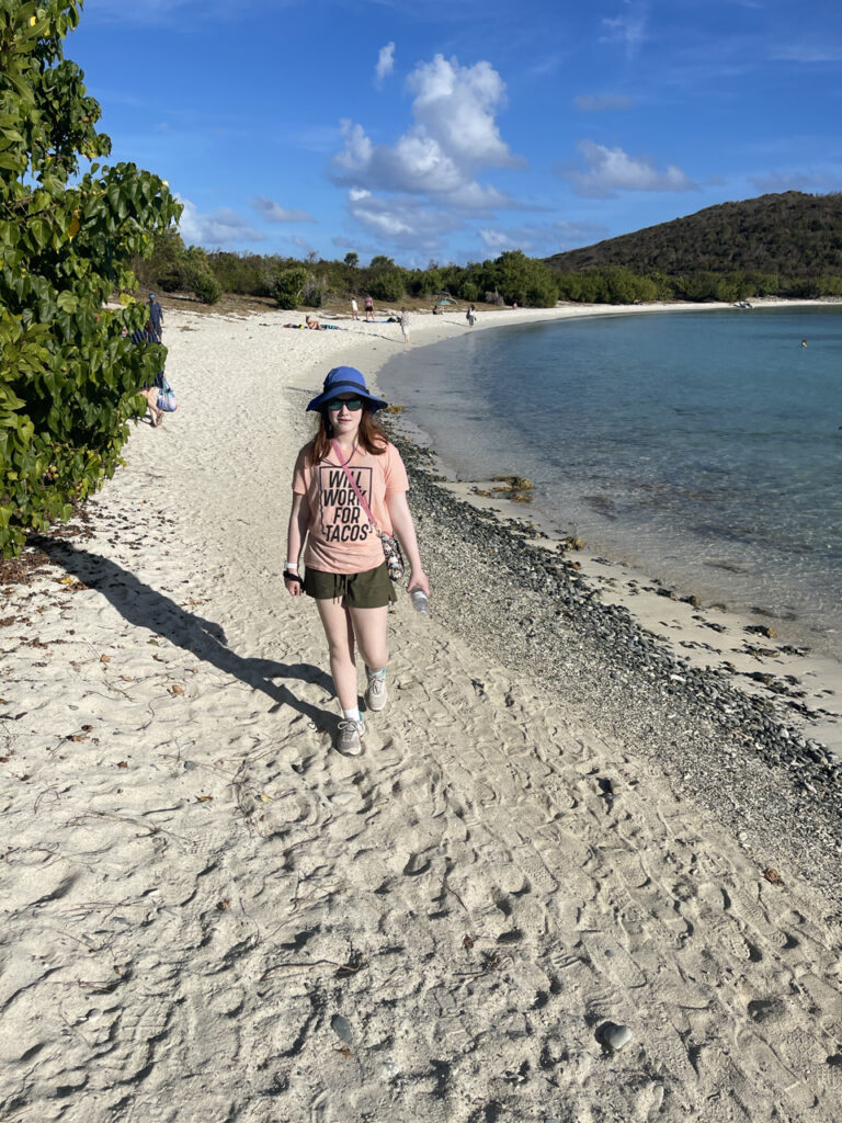 Cameron taking a stroll down Salt Pond Beach wearing shorts, t-shirt and a sun hat along with sunglasses on a bright blue day with a few clouds in the sky.