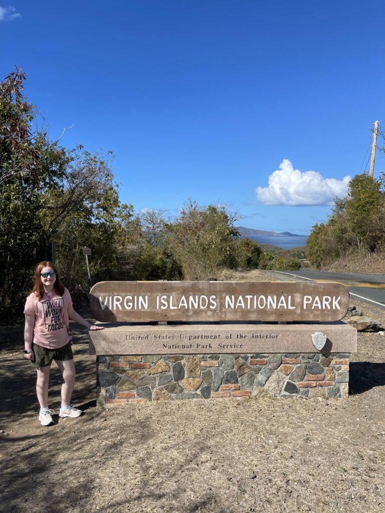 Cameron my red headed daughter, wearing shorts, t-shirt and sunglasses standing next to Virgin Islands National Park sign. The sky has only a few white puffy clouds on a spring day.