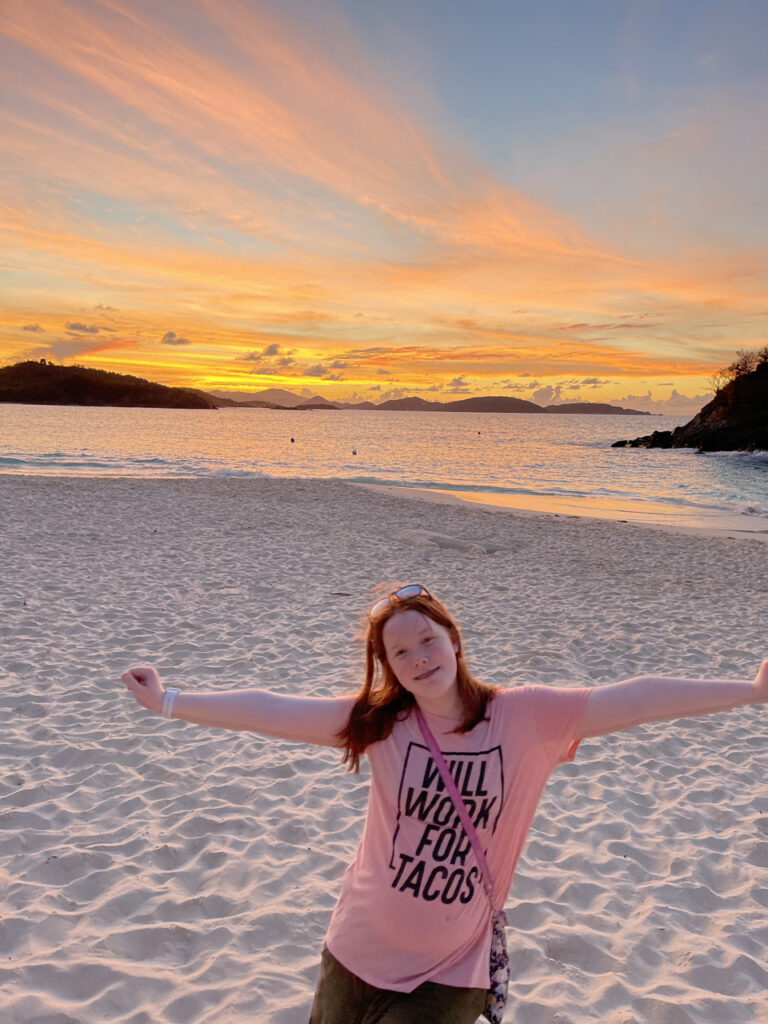 Cameron standing with her arms on in a t-shirt on Trunk Bay Beach in the Virgin Islands - the sky is fulled of bright yellow and orange clouds that are being reflected down into the water. 