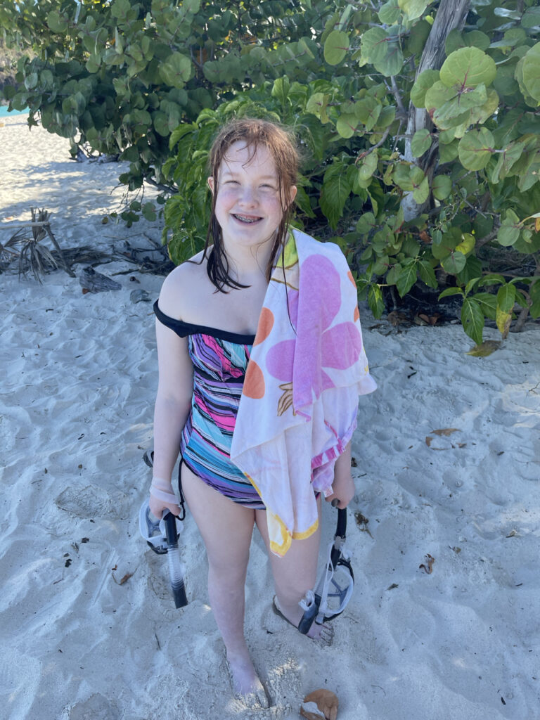 Cameron standing on the beach, wet from the water, in her bathing suit and towel - holding both of our Snorkel gear at Trunk Bay Beach.