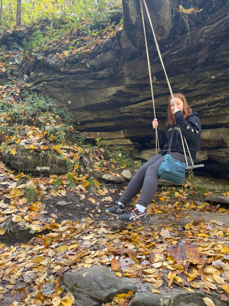 Cameron on a rope swing near Dunloup Creek with fall leaves on the ground and a large rock wall behind her.