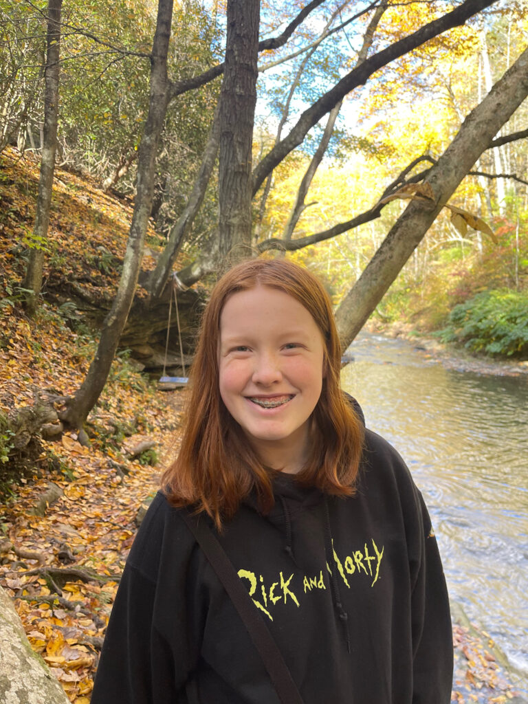 Cameron standing on the shores of the Dunloup Creek at the height of fall colors, with leaves on the ground and on the trees. 