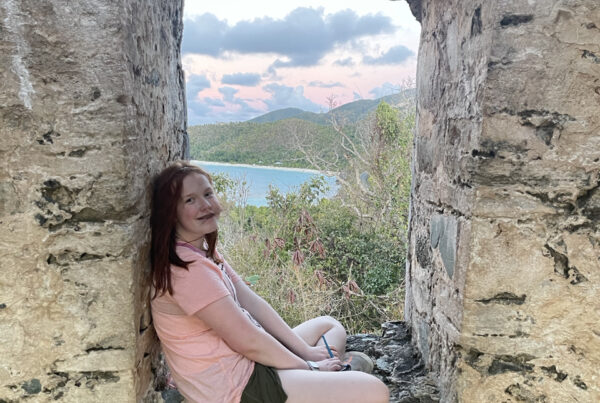 Cameron sitting in the window, legs crossed of what remains of the Sugar Mill - the ocean and clouds can be seen in the background.