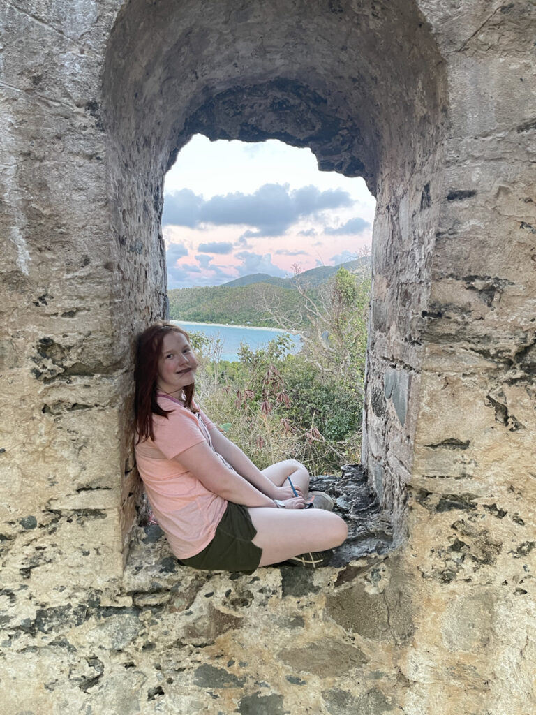 Cameron sitting in the window, legs crossed of what remains of the Sugar Mill - the ocean and clouds can be seen in the background.