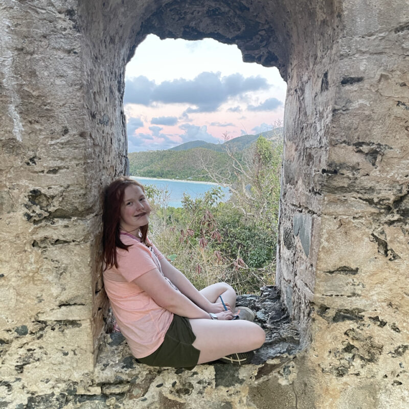 Cameron sitting in the window, legs crossed of what remains of the Sugar Mill - the ocean and clouds can be seen in the background.