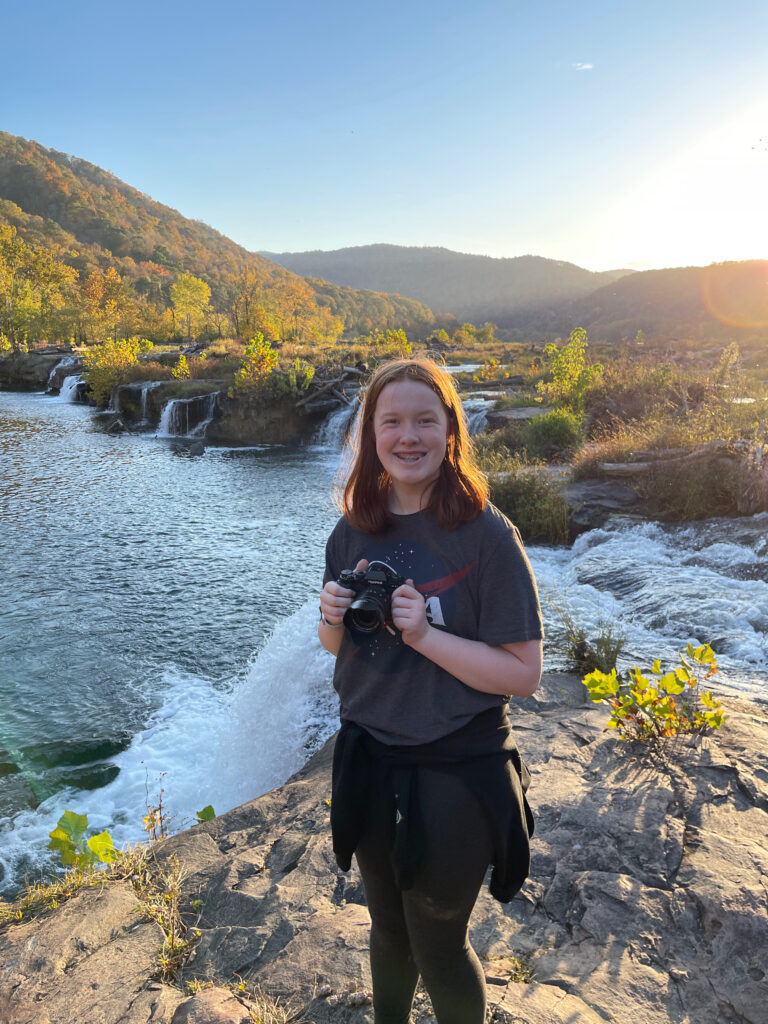 Cameron smiling and holding her camera standing on rocks on top of the waterfalls at Sandstone Falls in New River George National Park. The late afternoon light is just hitting the mountains in the distance and all the fall colors have come to life.