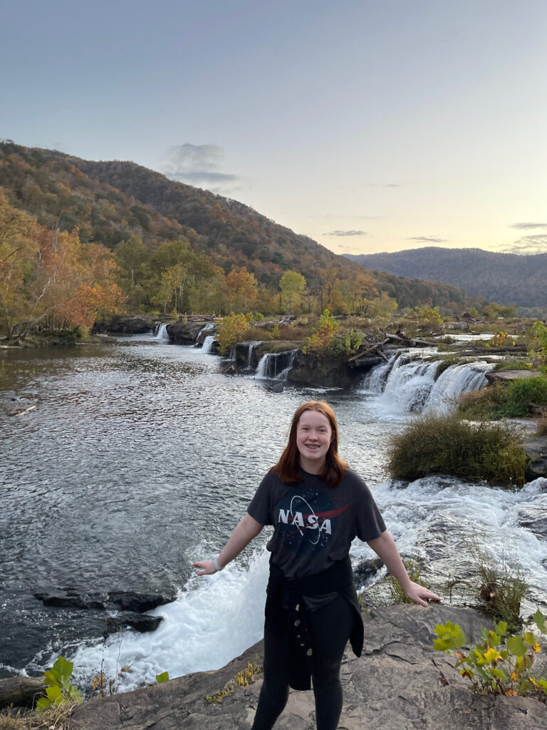 Cameron out at the edge of the rocks by the water fall at Sandstone Falls on the new river. She is all smiles and behind her is rushing water and a stunning sunset sky.