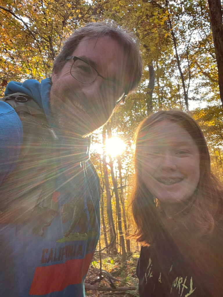 Cameron and I on a hiking trail in New River George - the trees are yellow with fall colors and the sun is just breaking through between the trees and us as we look down at my camera. 