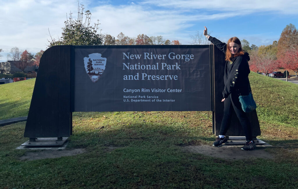 Cameron standing next to the New River Gorge National Park sign holding her arms up to frame the sign, wearing pants and a hoodie. 