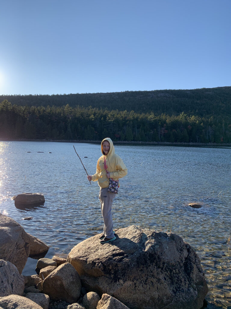 Cameron standing on a massive boulder holding a stick on the shores of Jordan Lake in Acadia National Park.