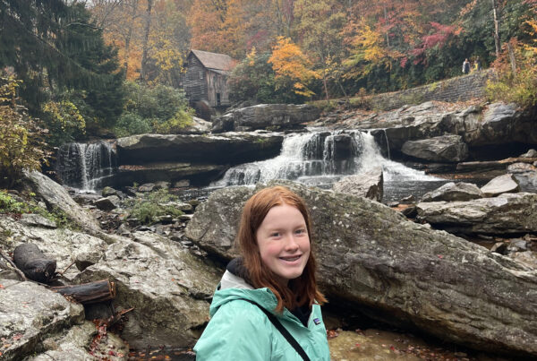 Cameron wearing a rain jacket, standing in the rain, holding her camera in the middle of the river looking up at the Glade Creek Grist Mill.