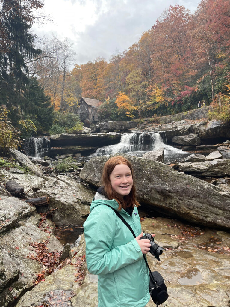 Cameron wearing a rain jacket, standing in the rain, holding her camera in the middle of the river looking up at the Glade Creek Grist Mill.