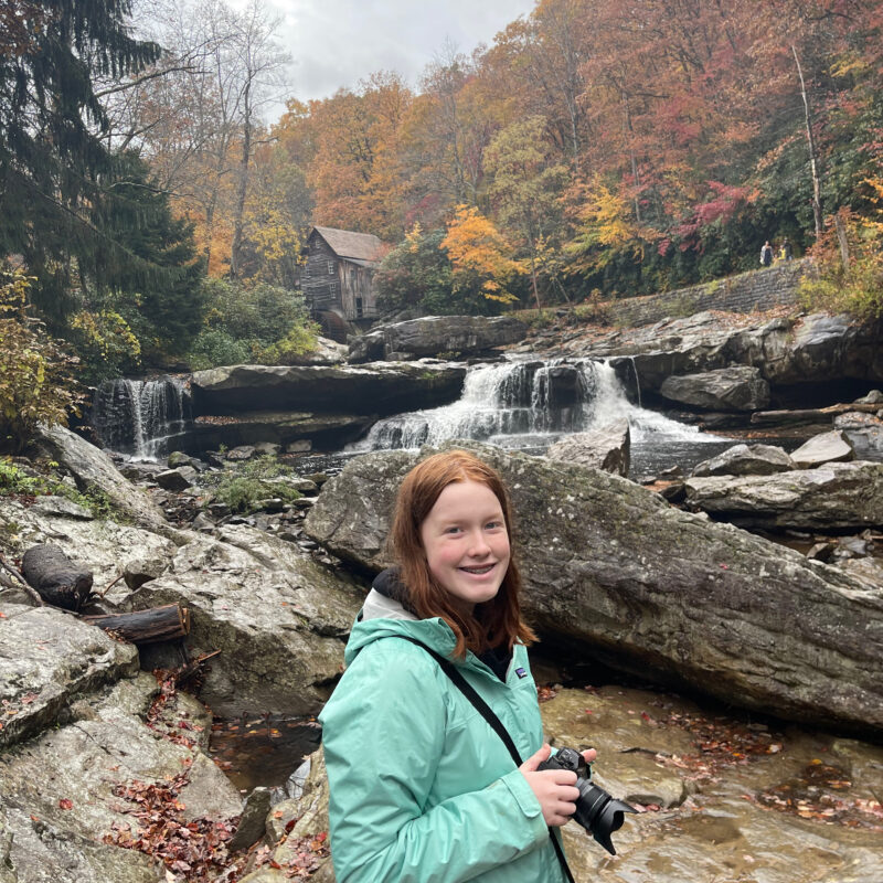 Cameron wearing a rain jacket, standing in the rain, holding her camera in the middle of the river looking up at the Glade Creek Grist Mill.