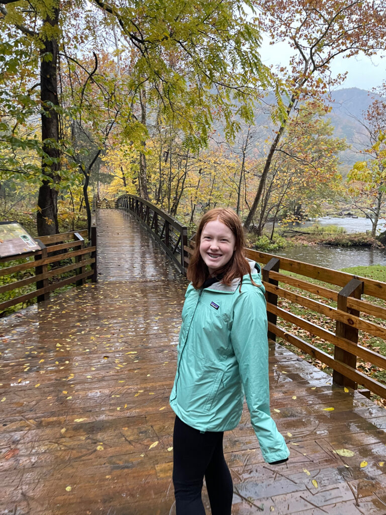 Cameron standing in her blue raincoat on the very wet boardwalk at Sandstone Falls on the New River. The trees are full of fall color and the rain is coming down.