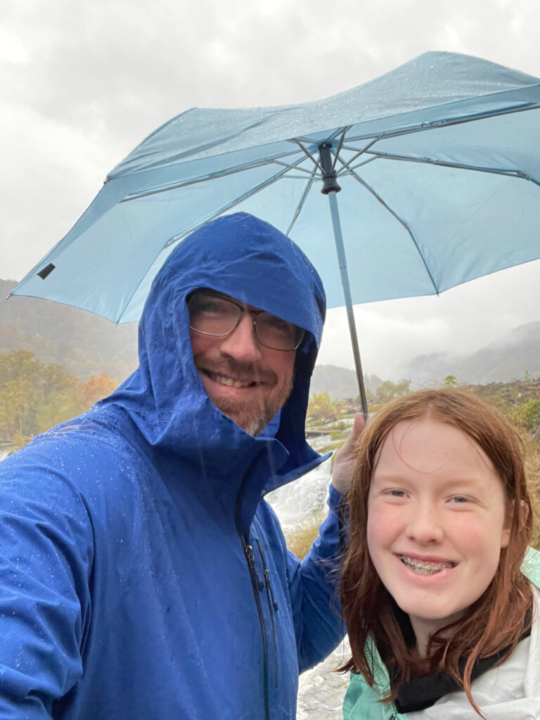 Cameron and I, both wearing rain coasts and holding an umbrella in the pouring rain at Sandstone Falls on the New River. Low clouds can be seen almost at ground level behind us on the mountains.