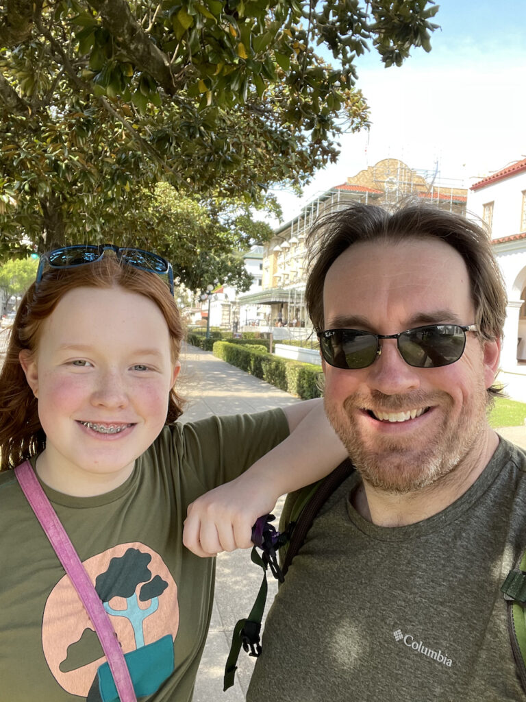 Cameron and myself both smiling in downtown hot springs, standing on the sidewalk with tress and shadows behind us. You can just see some of the old bathhouses. 