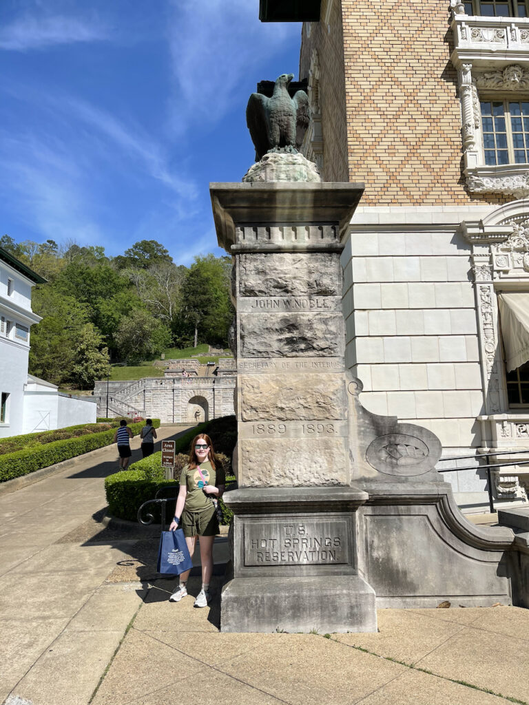 Cameron wearing sunglasses and carrying a bag from the visitors center. Standing next to a massive monument with an eagle on top and a tribute to John Noble inscribed on the stone. 