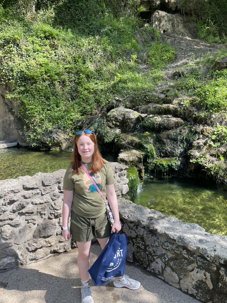 Cameron standing outside, holding a bag of goodies she just got from the visitors center. Behind her are man made pools to collect the hot spring water and behind them is the mountain with the water running down.