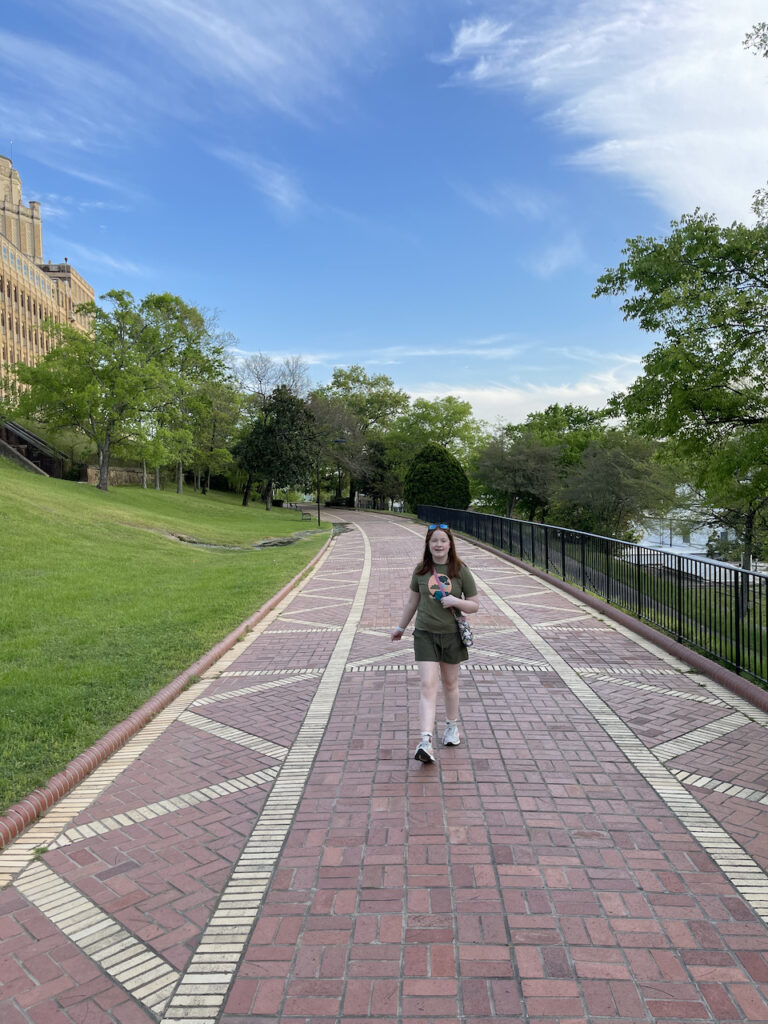 Cameron walking down the Grand Promenade in Hot Springs National Park. A big smile on her face and she walks over the impressive brick work on with blue skies and small white clouds above. 