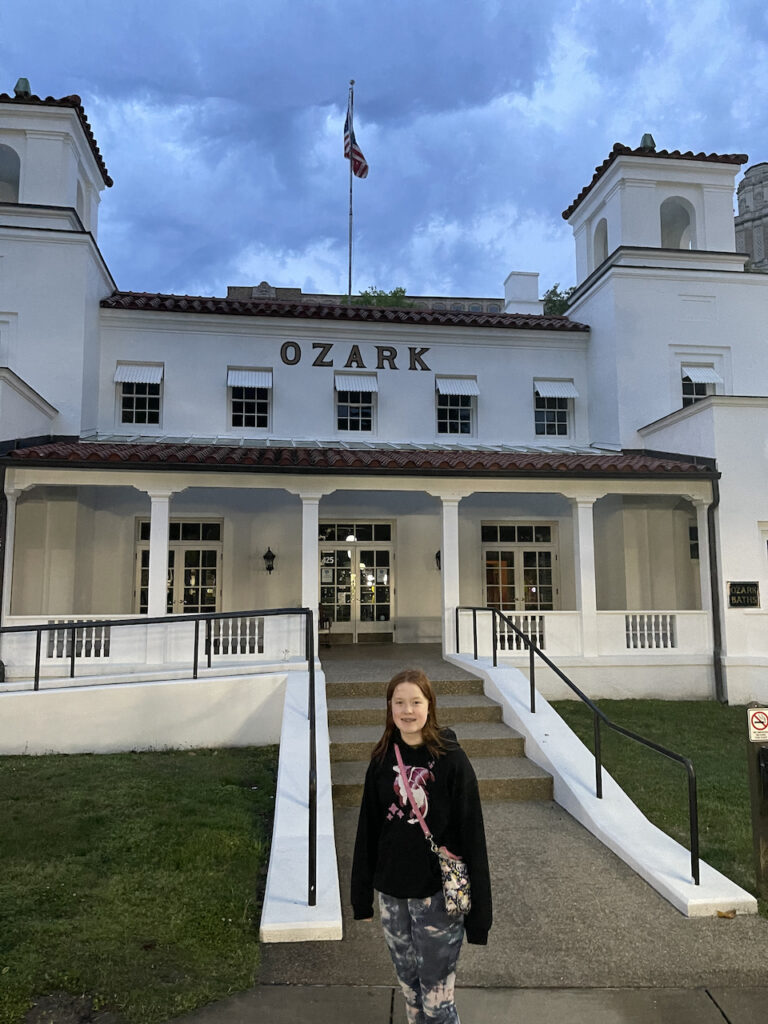 Cameron poses for a photo outside at the bottom of the stairs outside the Ozark bathhouse. Above an American flag flies and storm clouds loom overhead. 